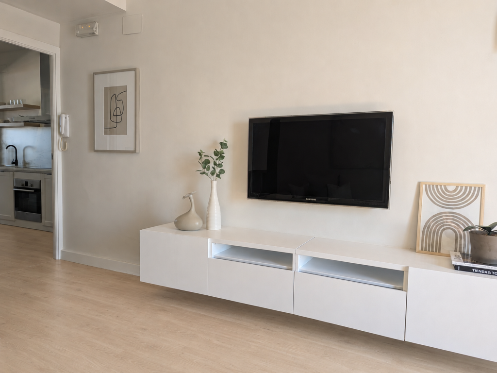 Televisión suspendida sobre mueble bajo blanco con jarrones, lámina geométrica y detalles vegetales en una pared clara